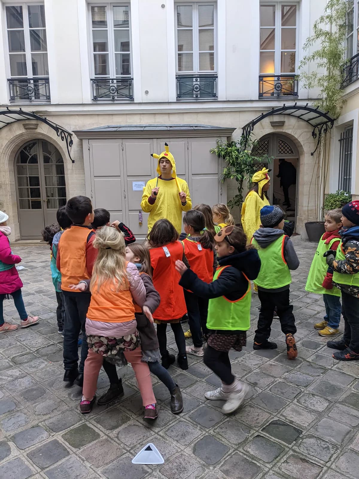 Une animatrice en costume jaune lance un jeu à des enfants en chasubles, dans la cour d’un immeuble