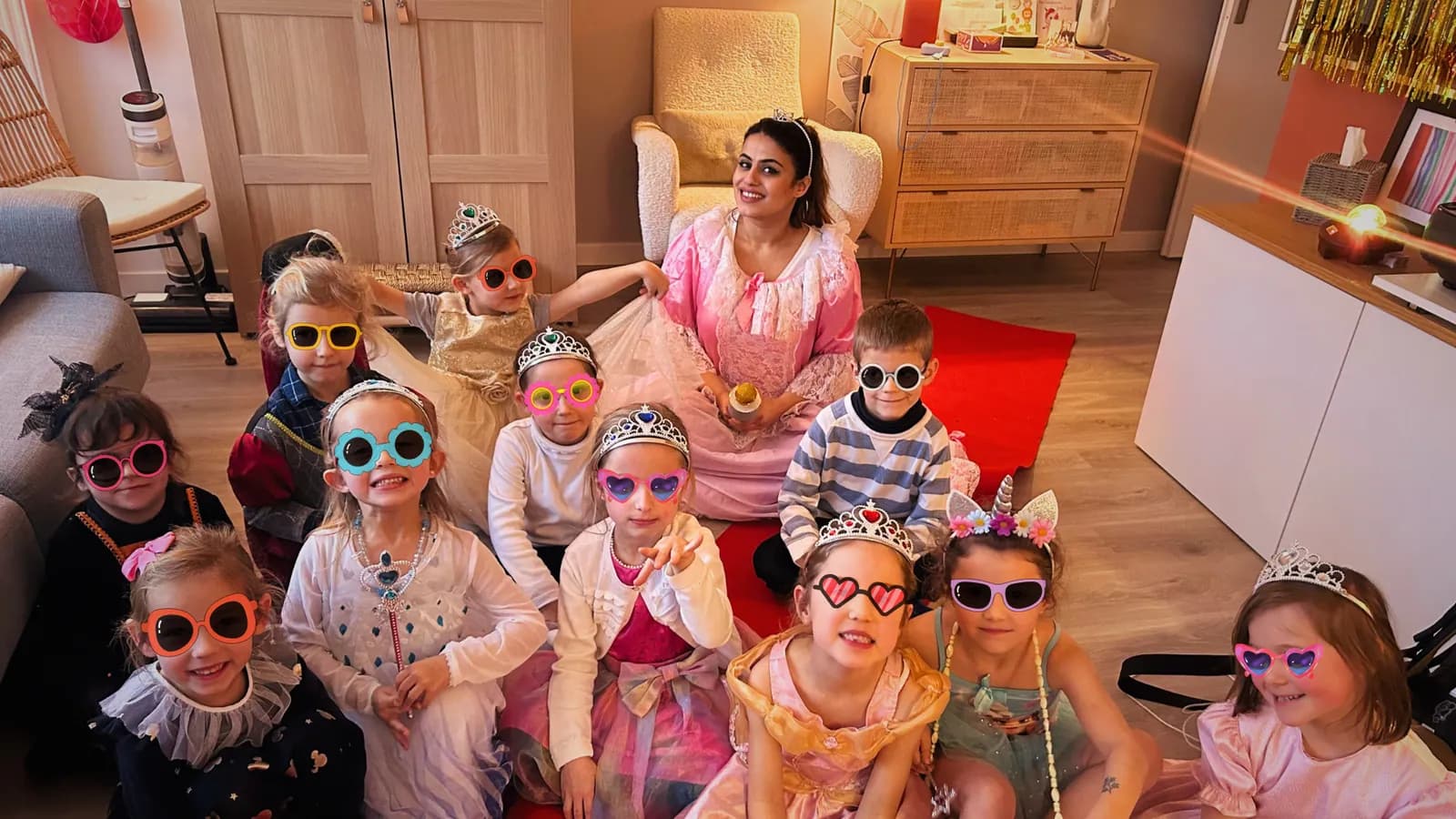 A host in a princess dress with a dozen children in tiaras and colourful sunglasses, in a living room