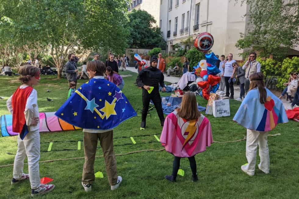 Enfants en capes de super-héros face à un animateur Kidays lors d’une animation en extérieur, parents autour