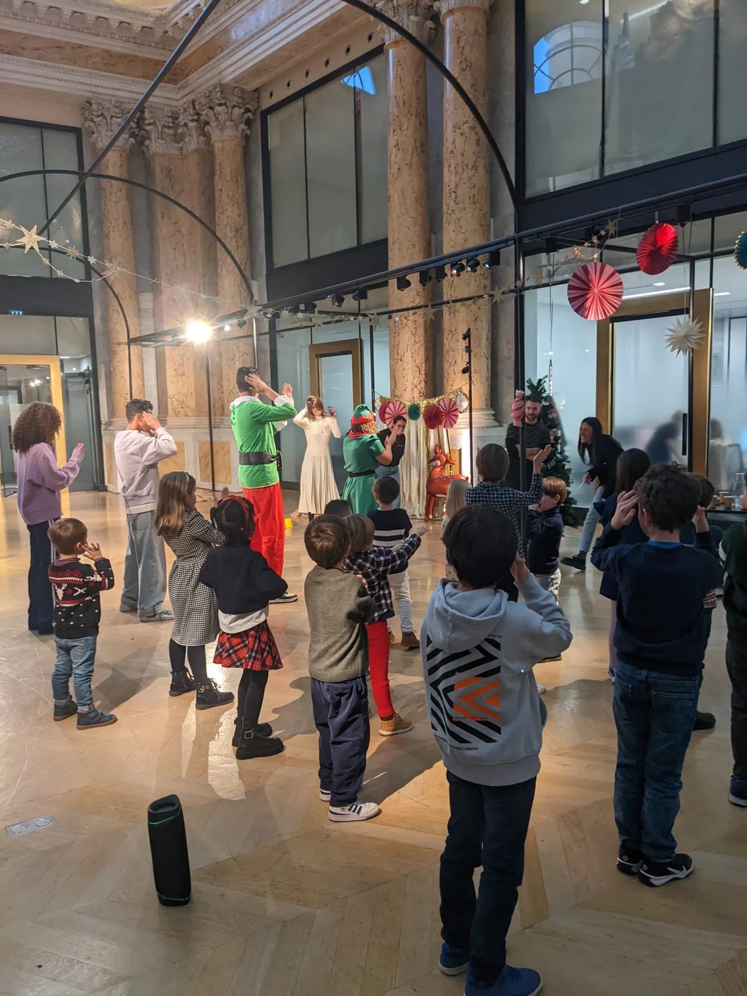 Kidays hosts in elf costumes leading a dance routine with children in a company lobby decorated for Christmas