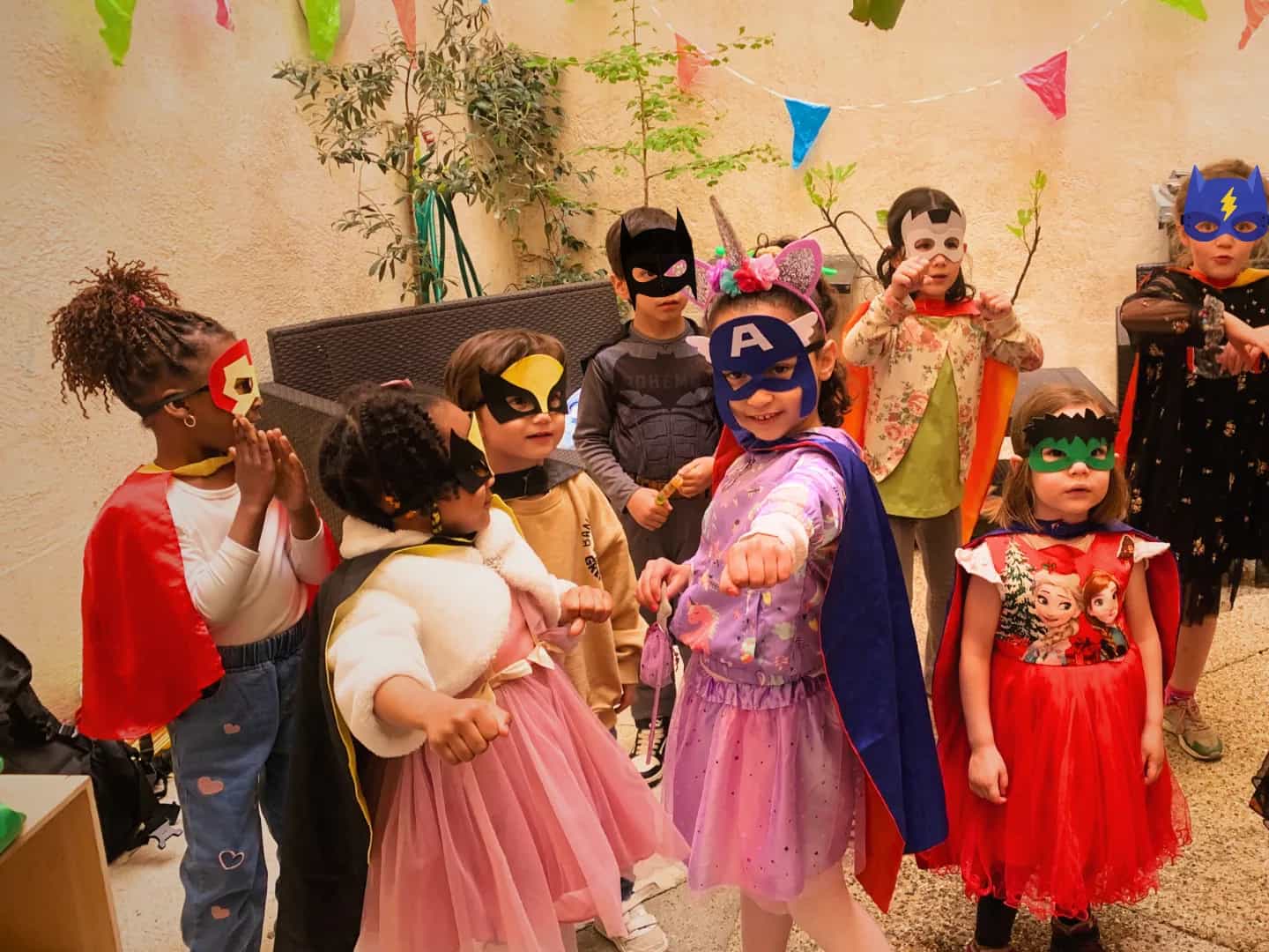 Young children in superhero capes and masks, fists out under a string of party bunting.