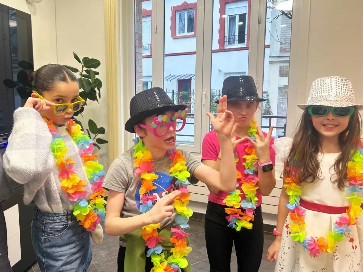 Four children in flower garlands, sequinned hats and novelty glasses, striking a pose at an indoor party.
