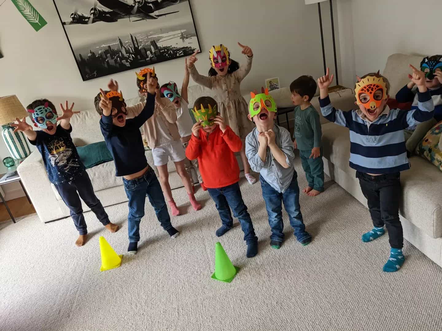 Des enfants en masques de dinosaures, bras levés, rugissent dans un salon, des plots de couleur posés sur le tapis.