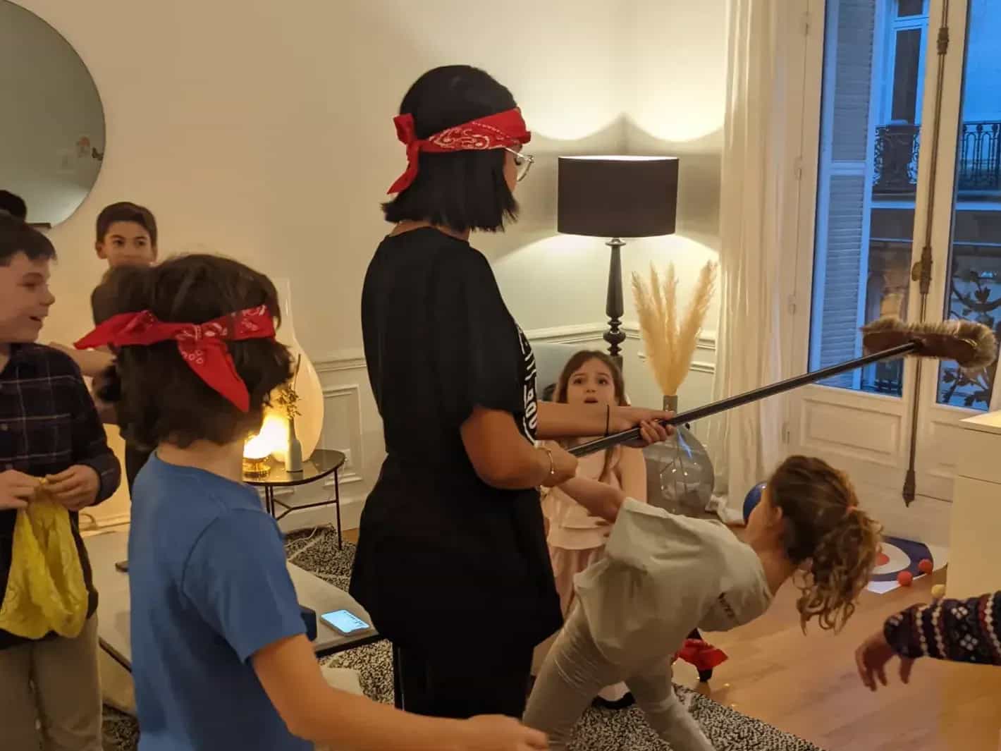 Une animatrice aux yeux bandés mène un jeu avec des enfants dans le salon d’un appartement.