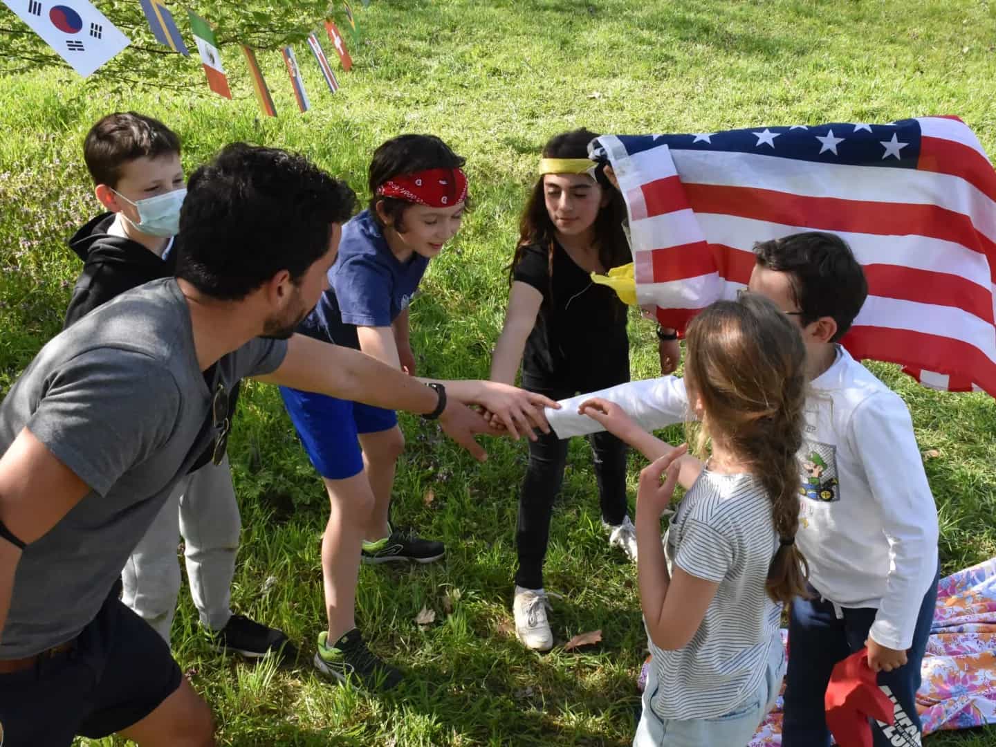 A host and a group of children join hands in the middle of the circle on a lawn, in front of a row of flags, before a team challenge.