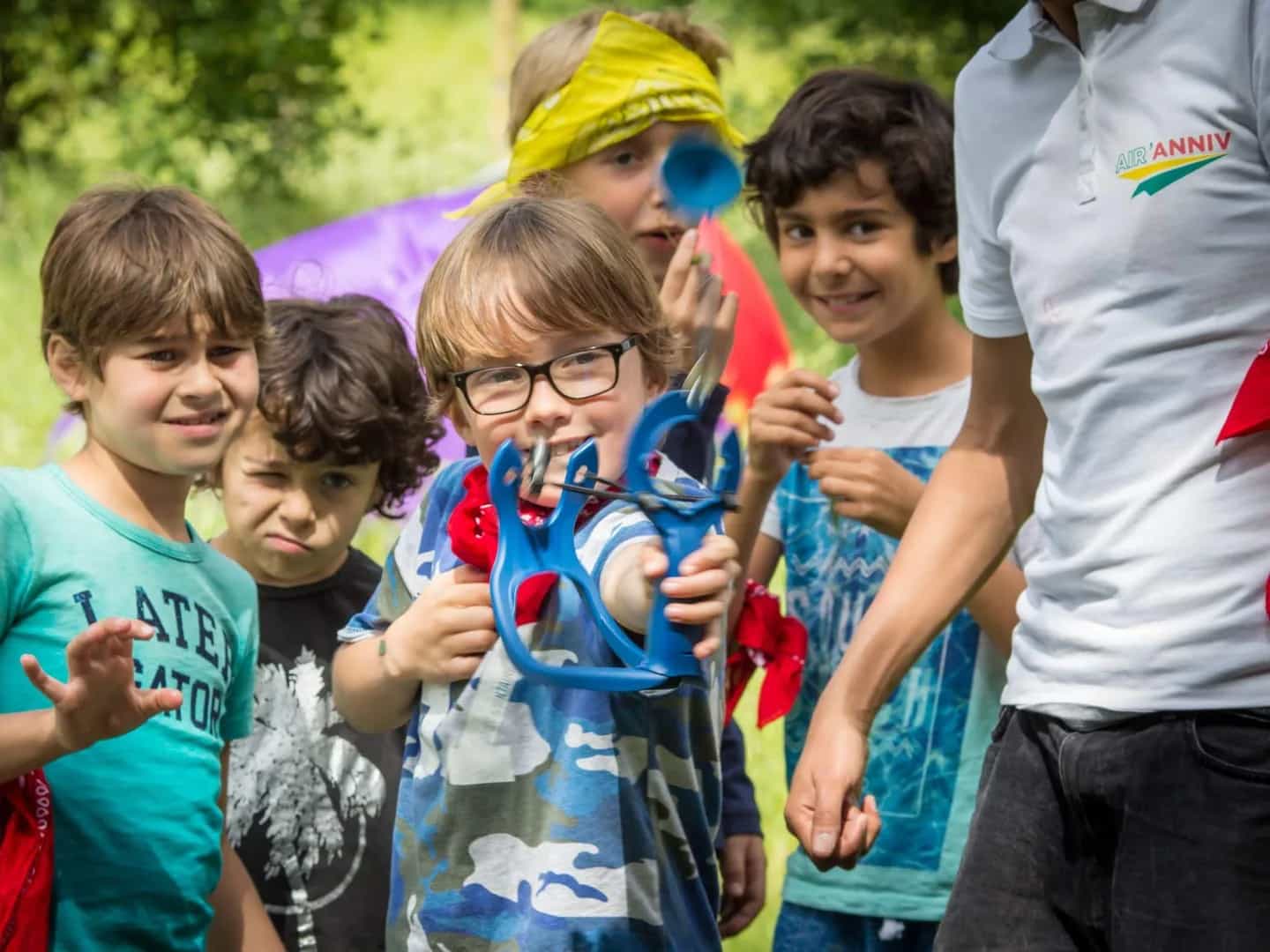 A group of children mid-game in a park, one of them holding a blue foam launcher out towards the camera.