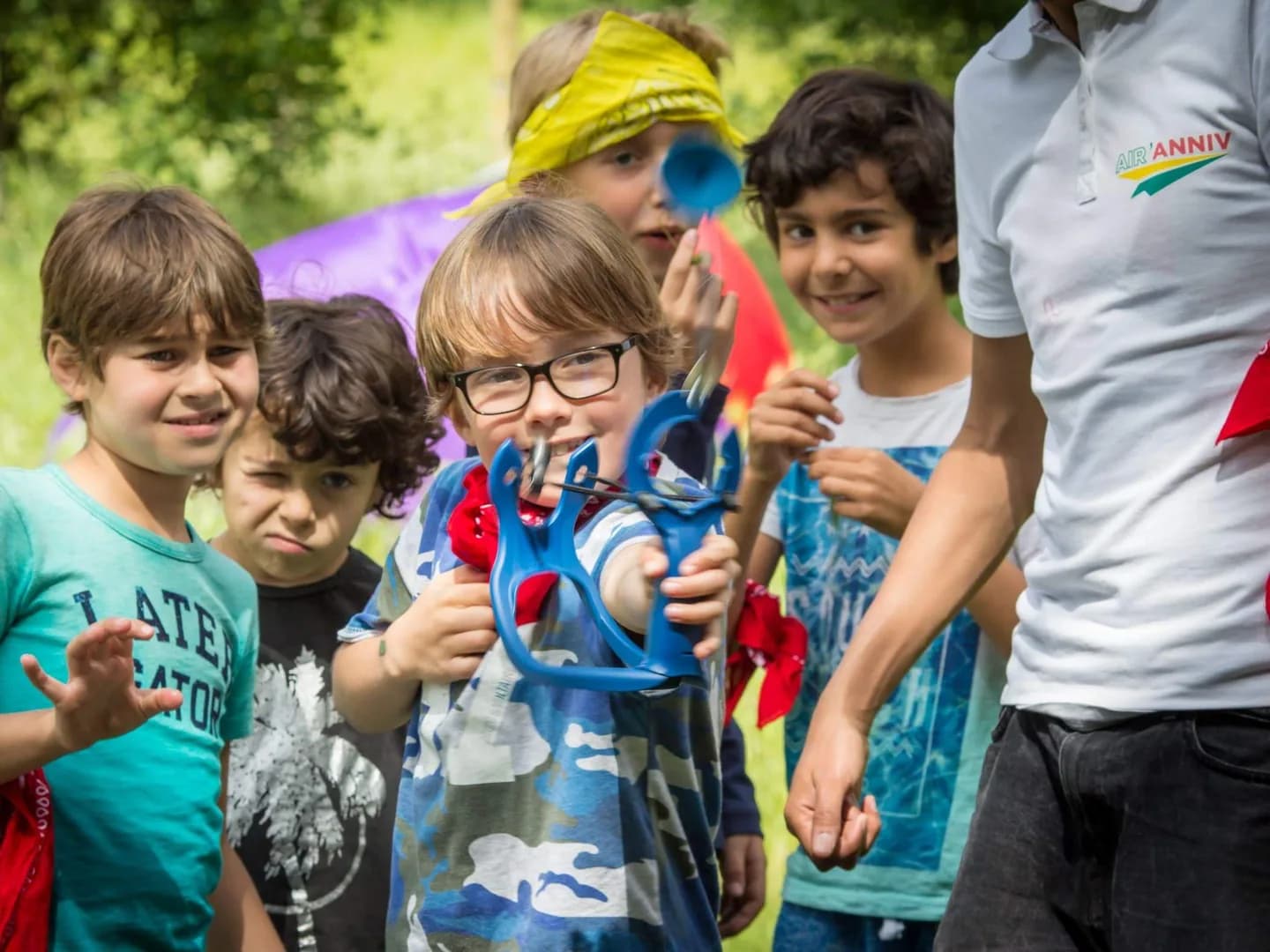 A group of children mid-game in a park, one of them holding a blue foam launcher out towards the camera.