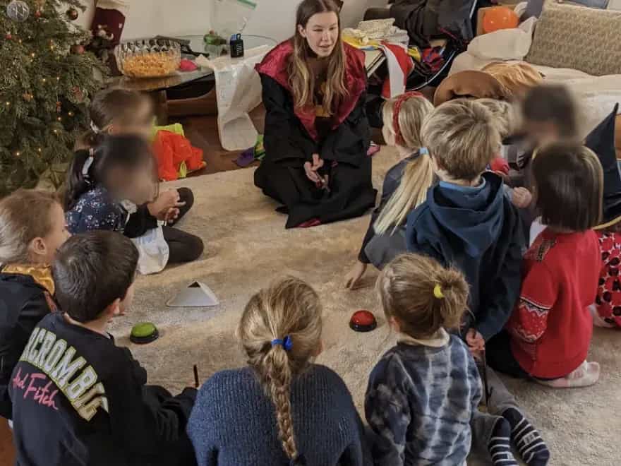 Une animatrice en robe de sorcier mène un jeu de buzzers avec des enfants assis en cercle sur le tapis du salon, à côté d’un sapin de Noël.