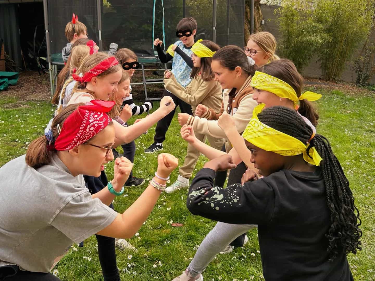 Two teams of children in red and yellow bandanas, fists raised and facing off in a garden during a challenge.