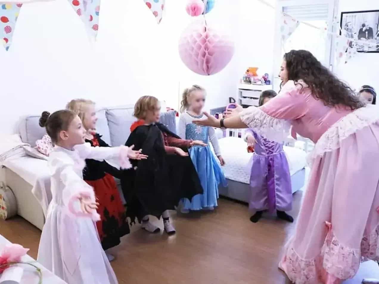 Une animatrice en robe de princesse rose fait danser des petites filles déguisées dans un salon décoré de fanions.